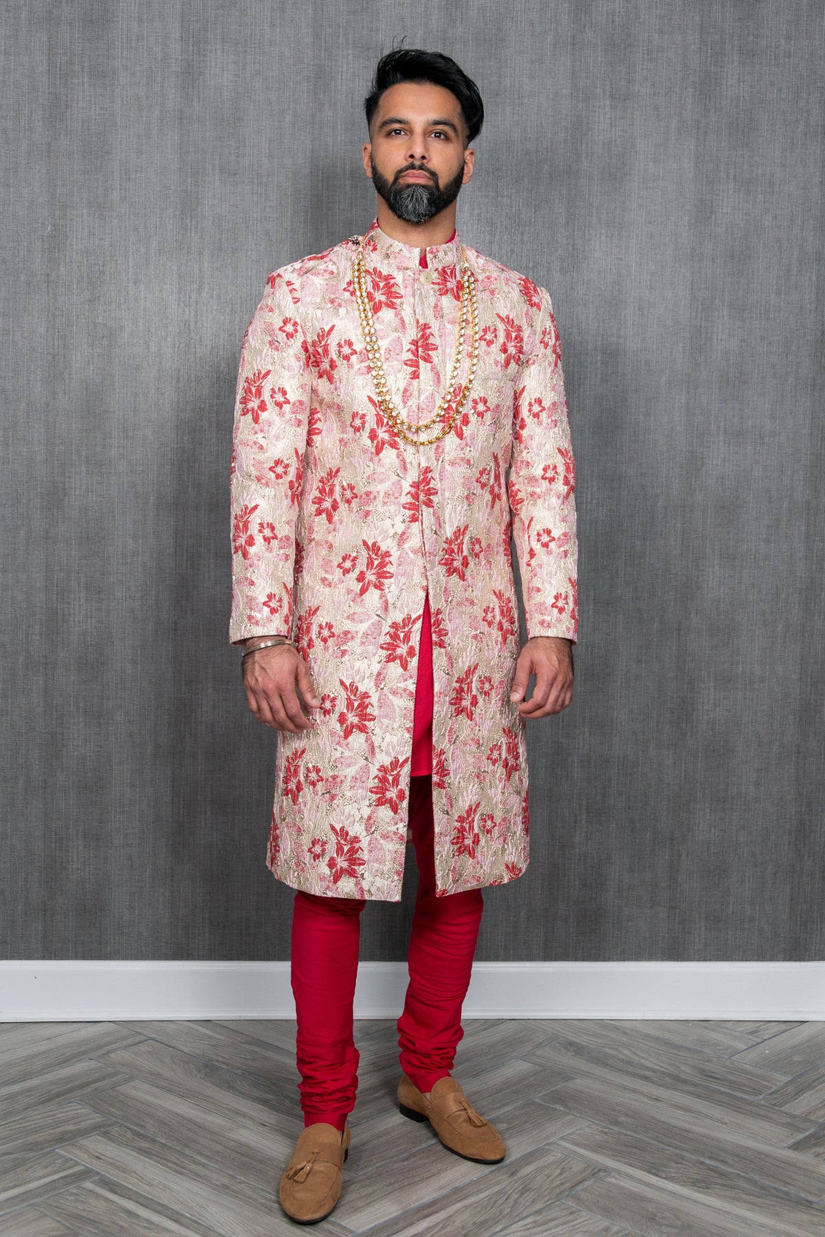 front view of man wearing AJAY red, cream, and gold sherwani with red pajama pants, brown loafers, and gold necklaces in front of a gray background