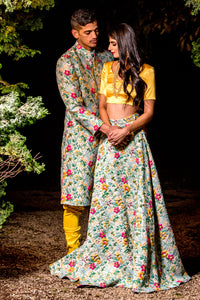 Front view- man wearing a multicolor Floral Jacquard Sherwani Jacket paired with yellow pajama pants holding hands with a woman wearing a matching lengha skirt and a satin yellow top. 