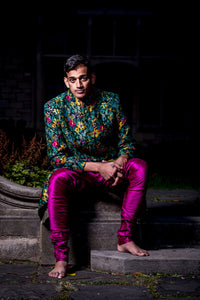 Front view- man wearing a dark green Floral Multicolor Sherwani Jacket paired with fuchsia pajama pants sitting in an outdoor setting 