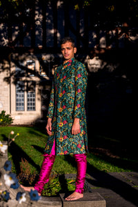 Side view- man wearing a Dark green Floral Sherwani Jacket with accents of yellow and red paired with fuchsia pajama pants standing in front of a shady outdoor background 