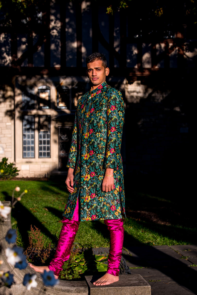 Side view- man wearing a Dark green Floral Sherwani Jacket with accents of yellow and red paired with fuchsia pajama pants standing in front of a shady outdoor background 