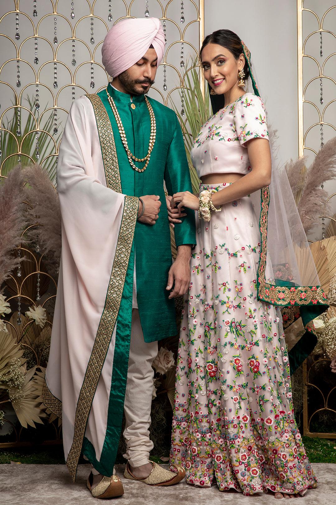 front view of a man wearing the ARUN silk sherwani jacket in evergreen with one gold button and wearing a cream stole with gold trim and cream pajama pants. Standing next to a woman in bridal set in front of a gold and floral background