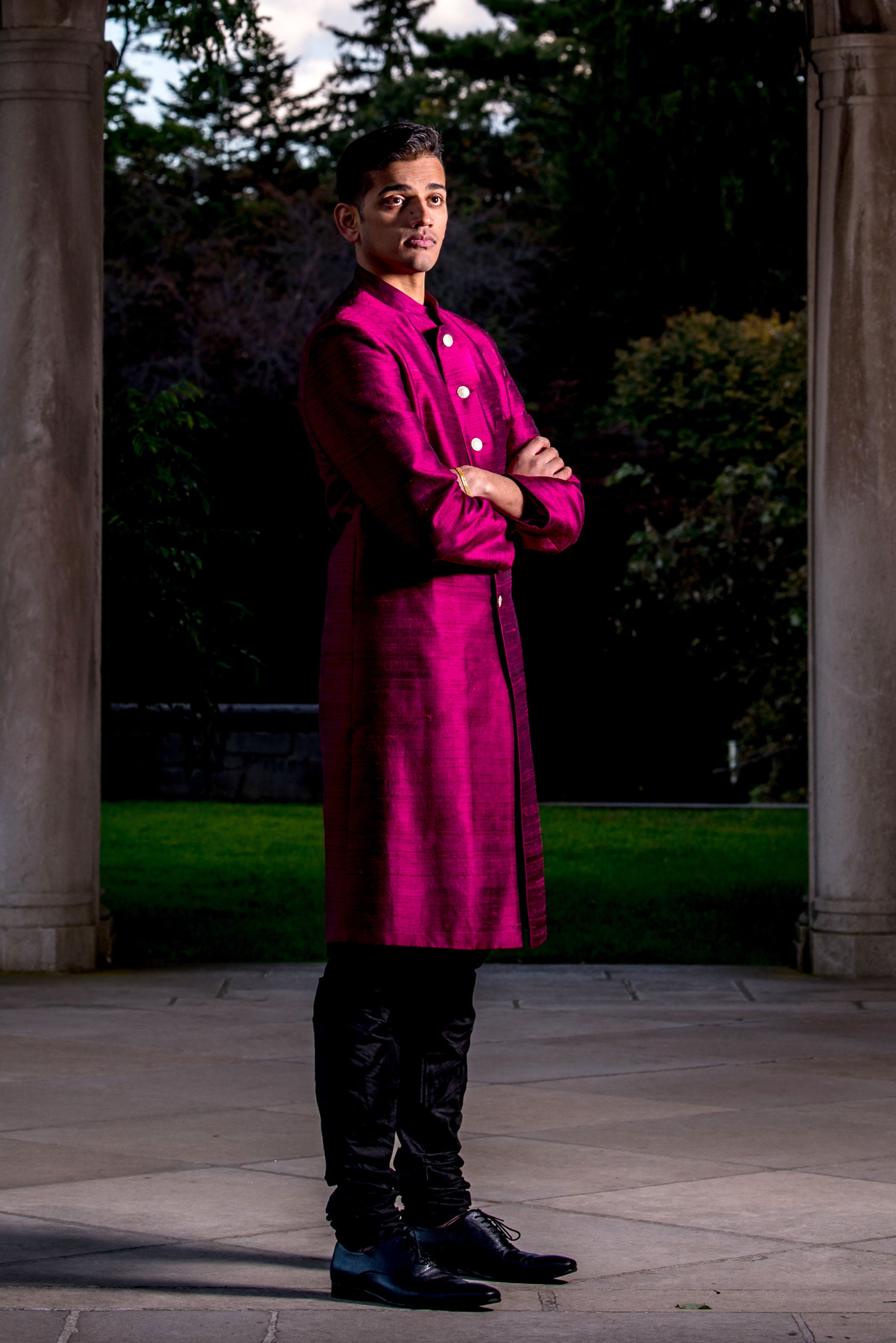 Side view- man wearing silk sherwani jacket in wine with black pajama pants and dress shoes in front of a forested background