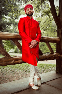 Front view- man wearing an Asymmetrical Sherwani Jacket in Red paired with cream pajama pants standing in front of a woodsy background 