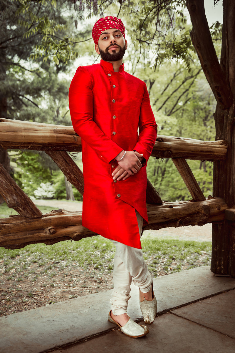 Front view- man wearing an Asymmetrical Sherwani Jacket in Red paired with cream pajama pants standing in front of a woodsy background 