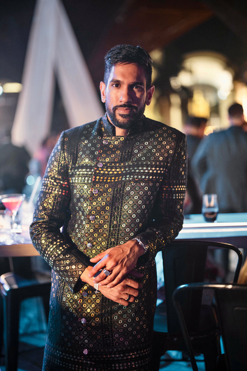Front view/upper body close up- Man wearing black colorful geometric sherwani in front of a bar setting