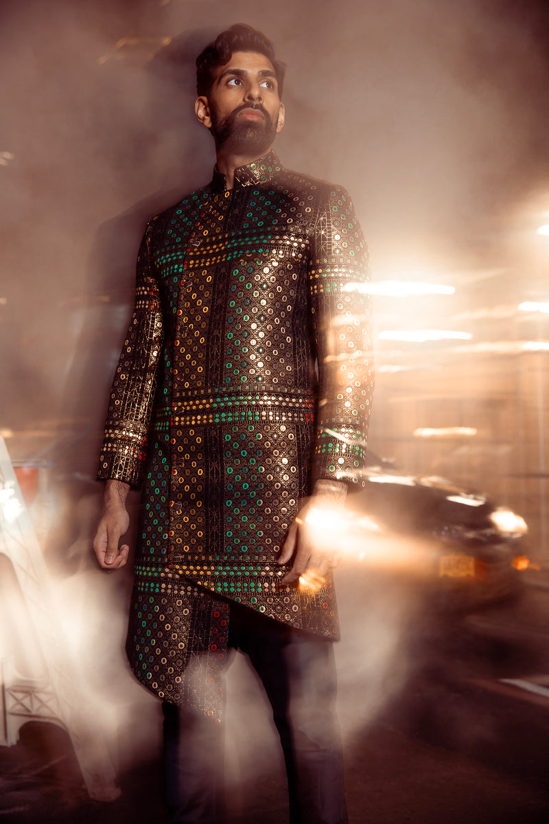 Low front view- man wearing a Geometric jacquard sherwani with gold and black in front of a blurry dark city background