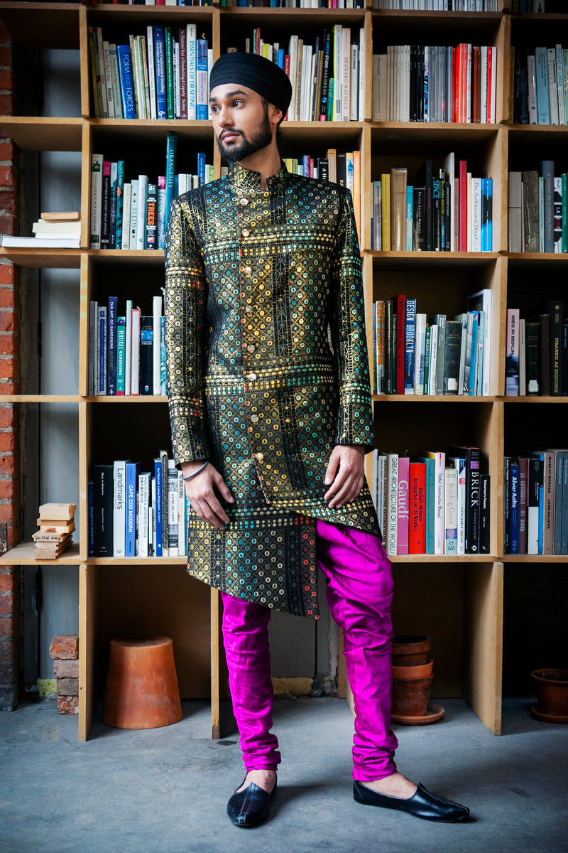 Front view- man wearing a black Geometric sherwani paired with fuschia pajama pants and a black turban, standing in front of a bookshelf looking to his right 