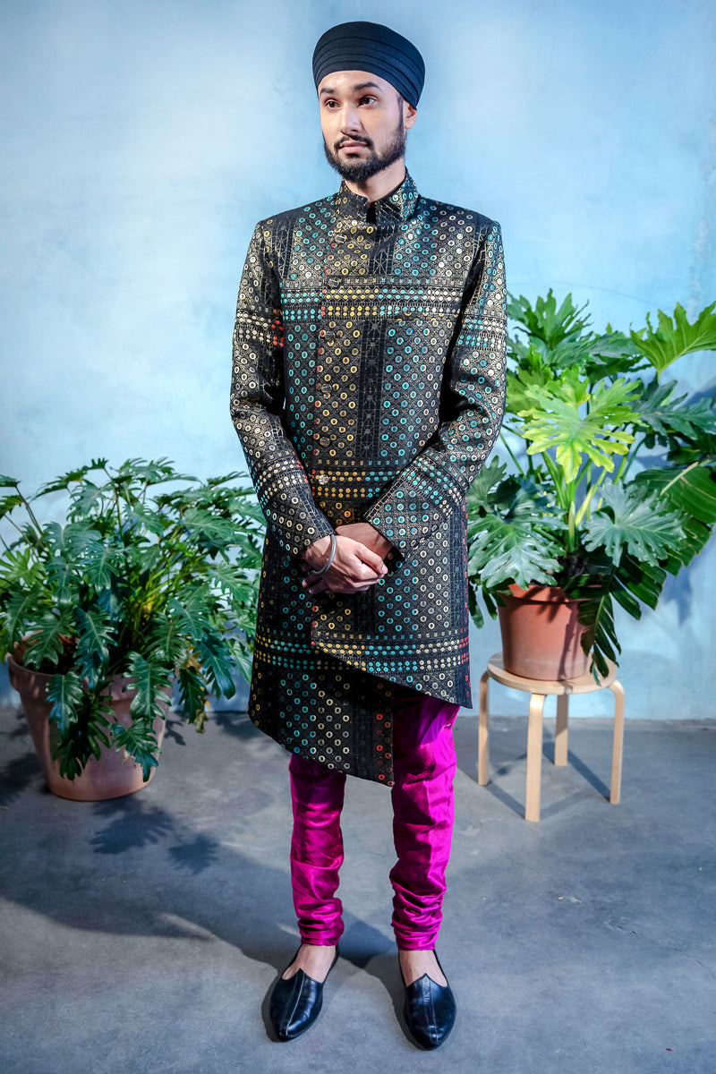 Front view- man standing wearing a black geometric sherwani jacket paired with fuchsia pajama pants and black shoes standing in front of a blue backdrop with plants on both his left and right side 
￼ 
