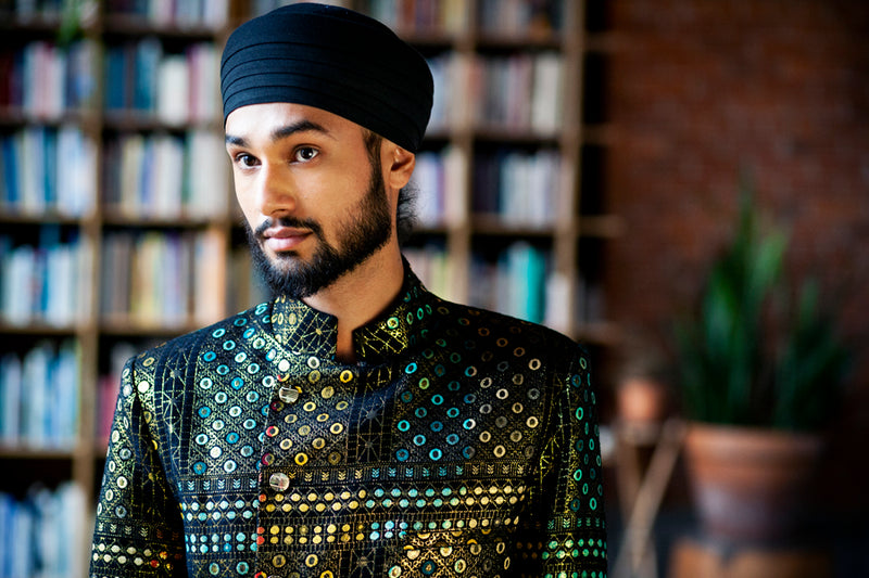 Close up side view- man wearing a black geometric sherwani jacket wearing a black turban in front of a blurry bookshelf background 
