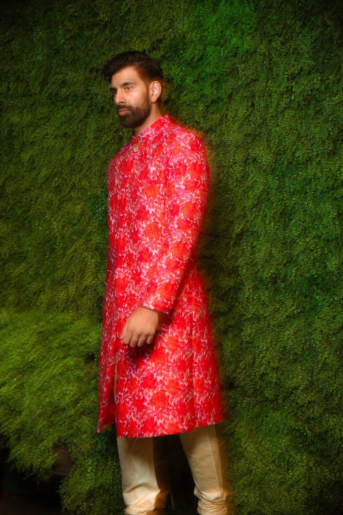 Side view- Man wearing red and pink floral sherwani in front of a grassy backdrop 