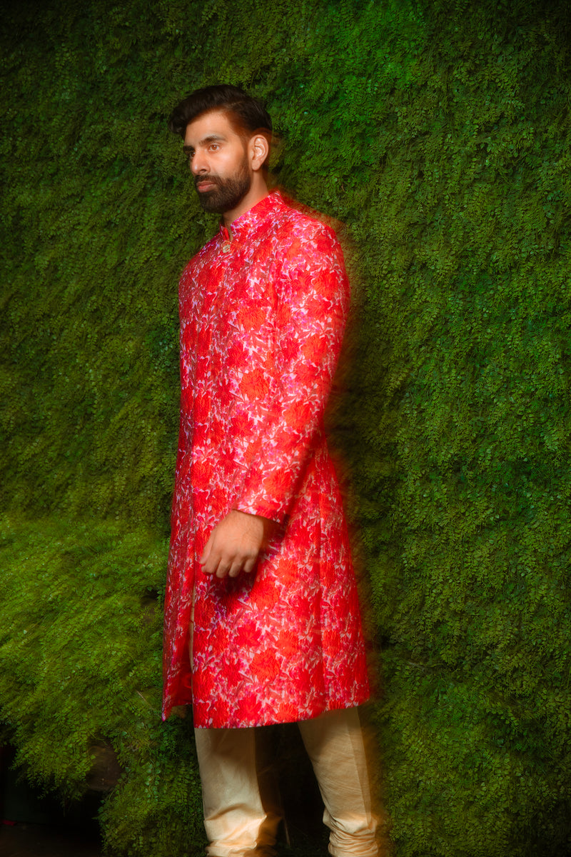 Side view- Man wearing red and pink floral sherwani in front of a grassy backdrop 