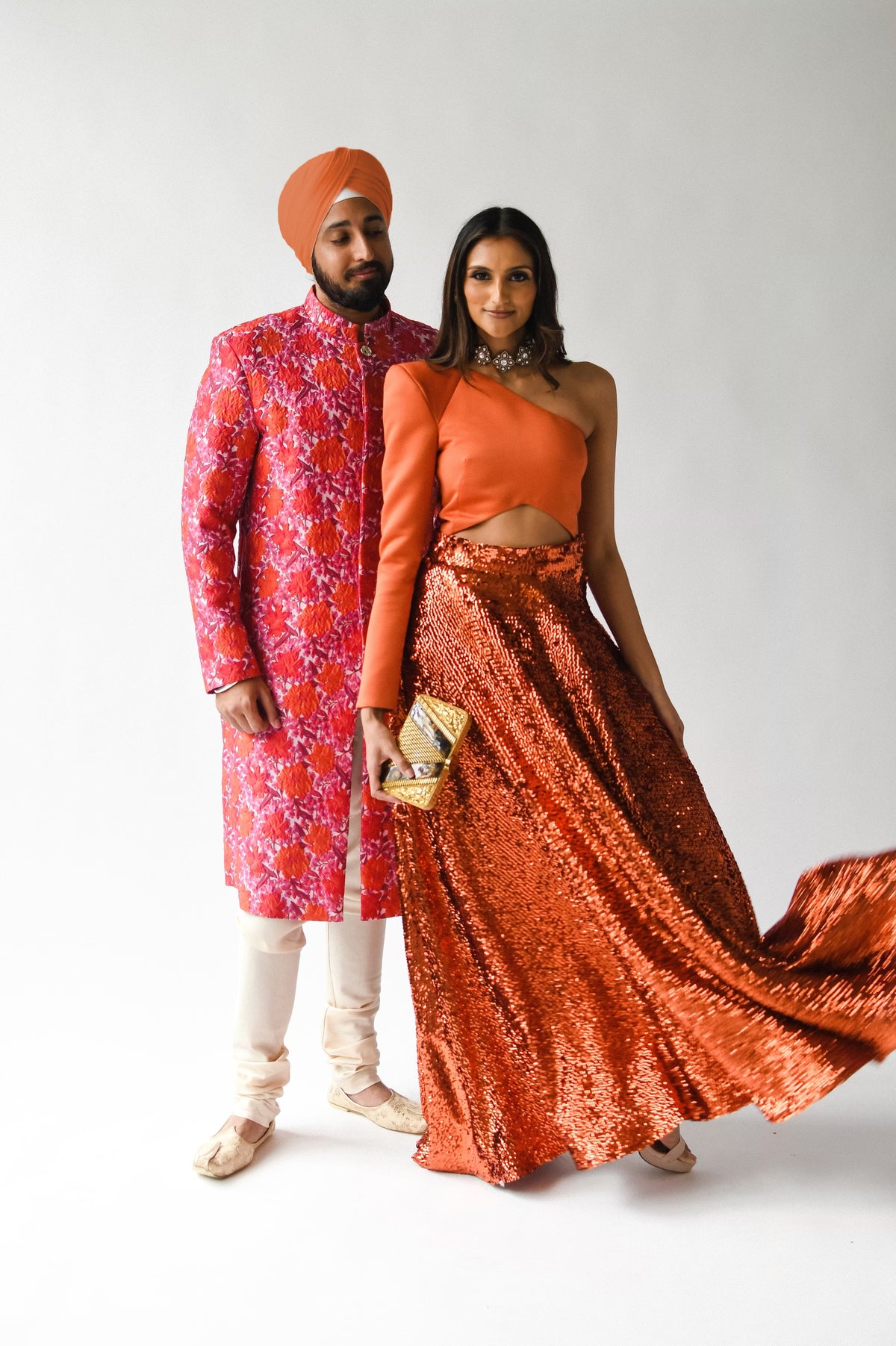 Front view- man wearing a red floral sherwani jacket standing next to a woman wearing a burnt orange lengha set standing in front of a white background 