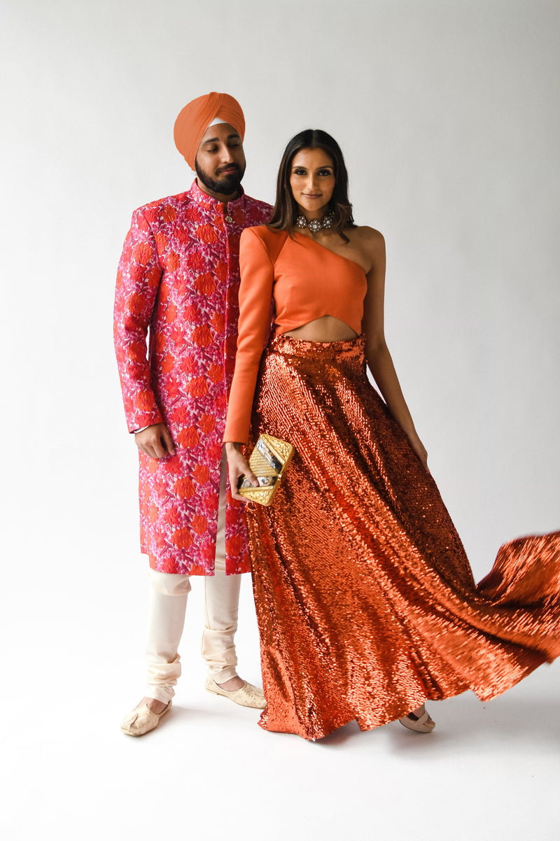 Front view- man wearing a red floral sherwani jacket standing next to a woman wearing a burnt orange lengha set standing in front of a white background 