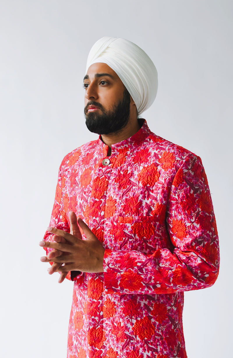 Side view- man wearing a red floral sherwani with a white turban standing in front of a white background 