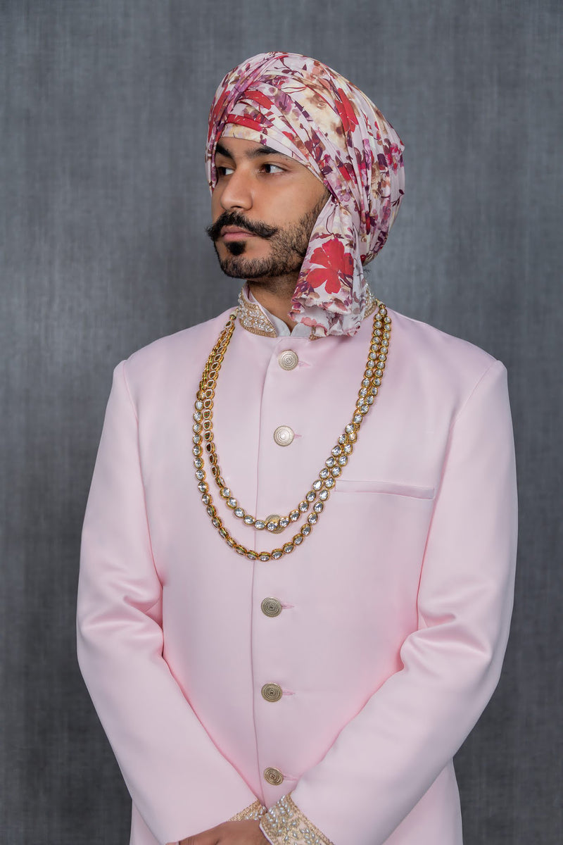 Close up view of PARAM pink sherwani jacket with pearl trim and a red floral turban. Man is wearing gold necklace in front of gray background
