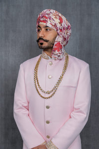 Close up view- man wearing a light pink sherwani jacket with pearl trim and gold buttons and wearing a white and red floral turban. Man is wearing two long gold chains. Man is standing in front of gray background