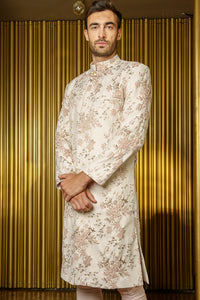 side view- man wearing PARK floral vines cream and pink sherwani with cream pajama pants in front of a golden background