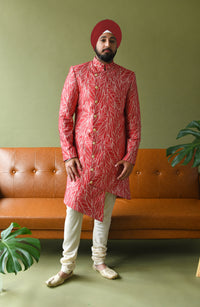 Front view- man wearing a red and white floral asymmetrical sherwani with white pajama pants standing in front of a green background with a brown couch 