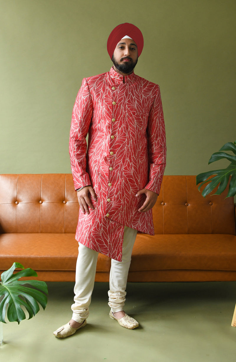 Front view- man wearing a red and white floral asymmetrical sherwani with white pajama pants standing in front of a green background with a brown couch 