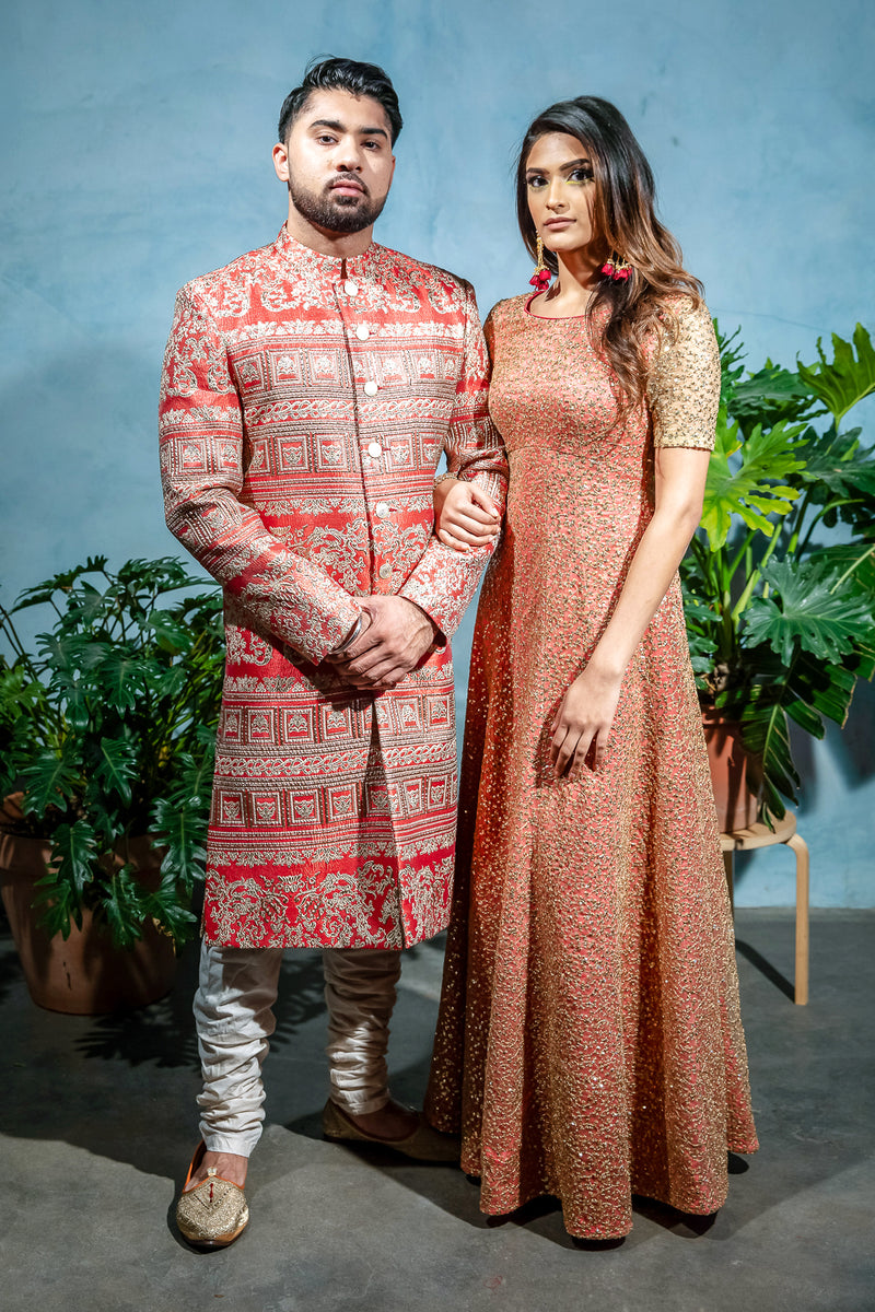 Front view- man wearing a red and gold Patterned Sherwani Jacket standing next to a woman wearing a coral dress with gold beading standing in front of a blue background 