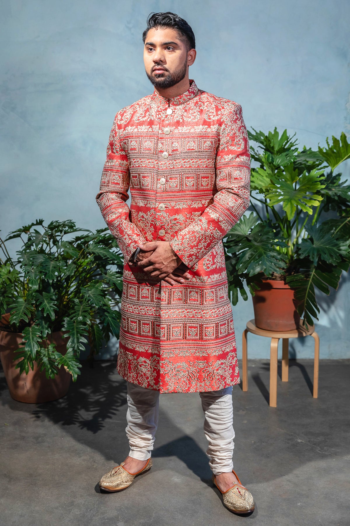 Drone view- man wearing a Sherwani Jacket in red and gold jacquard paired with white pajama pants standing in front of a blue background with plants 