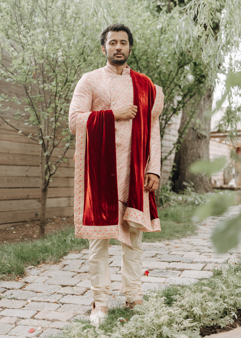 Front view- man wearing a red and gold striped sherwani with red velvet stole hanging off his left shoulder and right arm, in front of a forest like background 