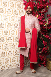 Front view- man wearing a Red and gold striped groomsman sherwani wearing red pajama pants, with a red velvet stole hanging over his right shoulder and left arm standing in front of a floral background