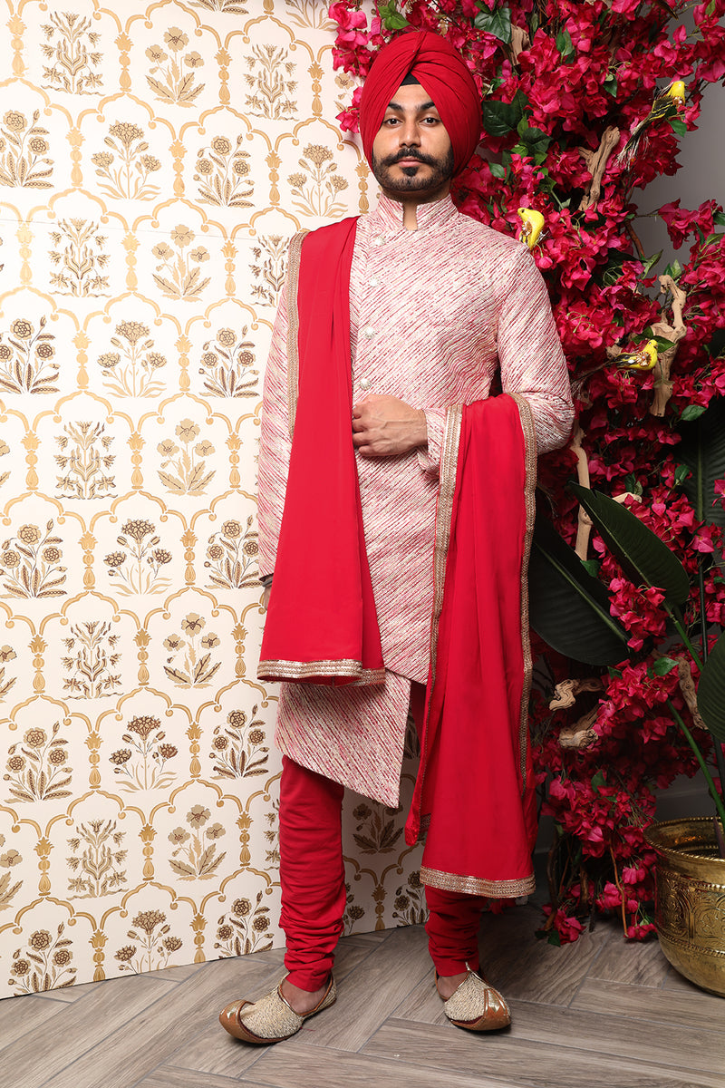 Front view- man wearing a Red and gold striped groomsman sherwani wearing red pajama pants, with a red velvet stole hanging over his right shoulder and left arm standing in front of a floral background
