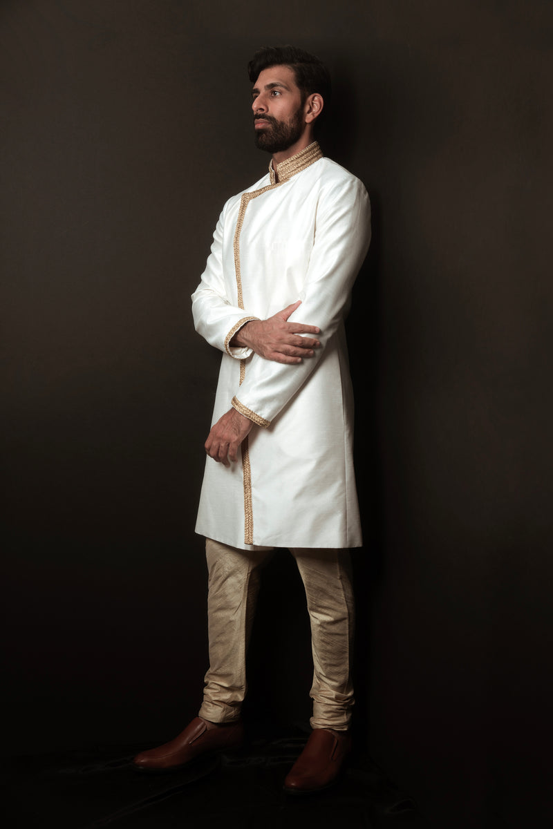 Groom with white sherwani with gold trim, gold pajama pants in front of a black background