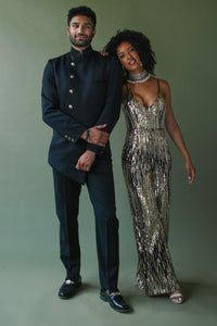 Front view- man wearing an asymmetrical black jacket with black dress pants standing next to a woman wearing a gold metallic jumpsuit, standing in front of a green backdrop 
