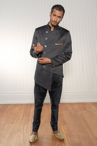 Front view- man wearing a Black asymmetrical jodhpuri jacket with square buttons paired with black pajama pants standing in front of a white background with wooden floors 
