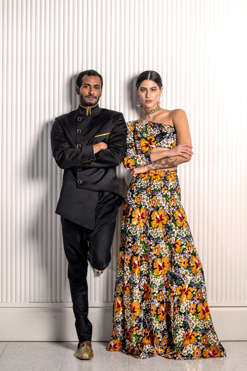 Front view- man wearing a Black asymmetrical jodhpuri jacket paired with black pajama pants standing next to a woman wearing a floral bridal sequin lehenga set, both posing in front of a white wall