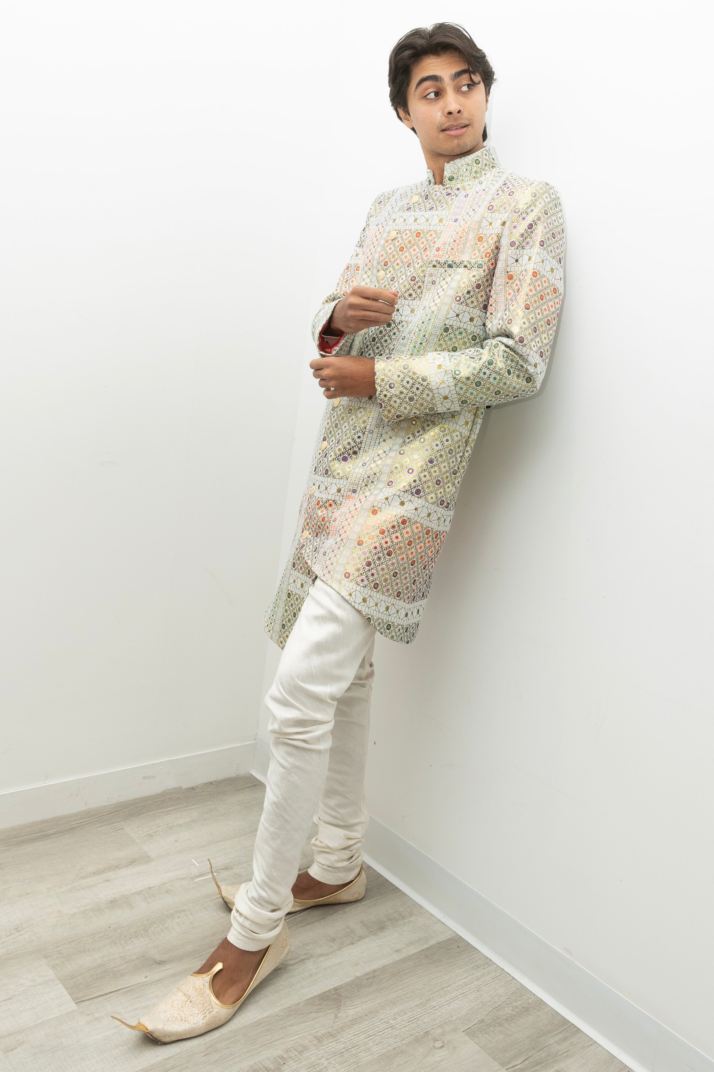 Side view- man wearing a white Geometric sherwani jacket with white pajama pants leaning on a plain white wall