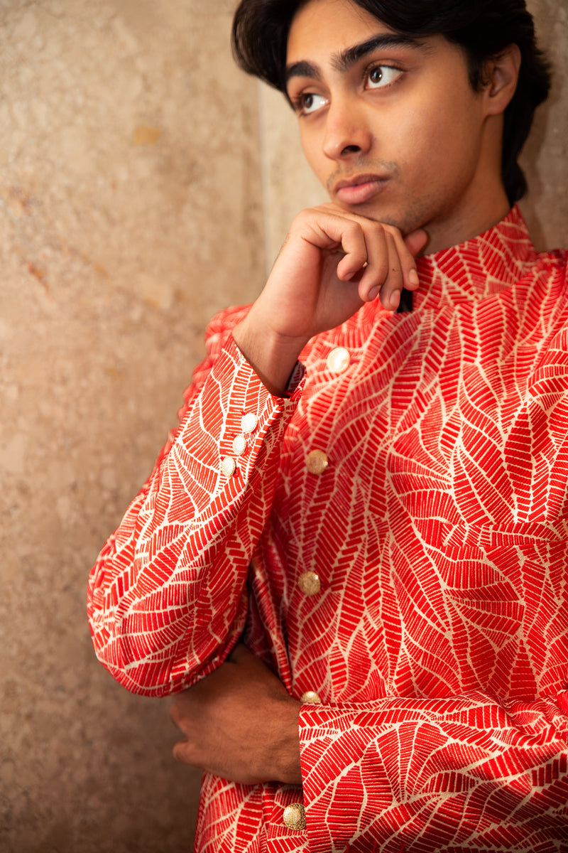 Close up side view- man wearing a red and cream floral sherwani with gold buttons posing on a tan wall corner  