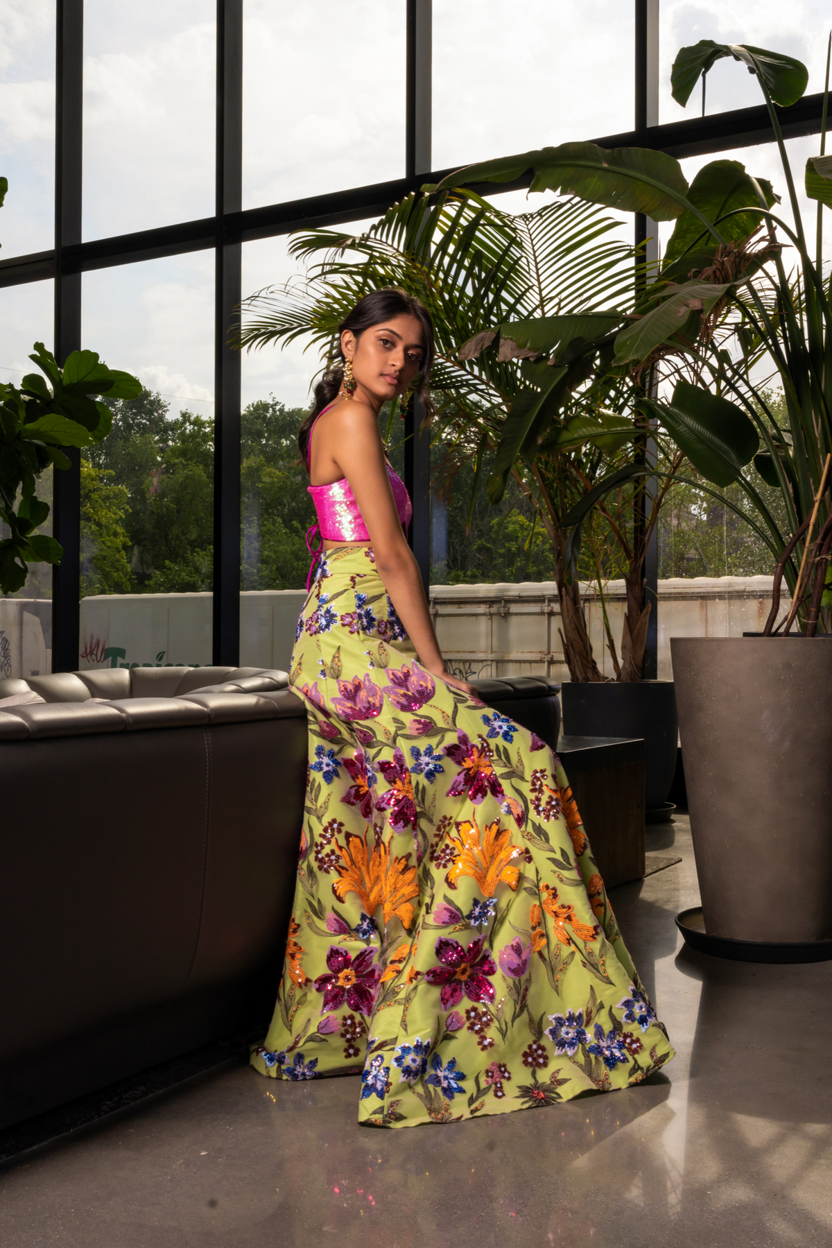 model leaning against a dark leather sofa in a high waisted lime green, floral sequin gabriella lehenga skirt. Styled with a neon iridescent pink sequin bralette top with a lace-up back. 